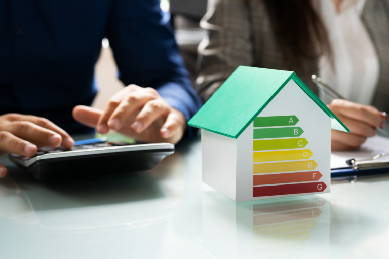 Dwójka specjalistów analizujących zużycie energii przy użyciu kalkulatora, obok makieta domu z kolorową skalą efektywności energetycznej od A do G – koncepcja świadectwa charakterystyki energetycznej budynku.