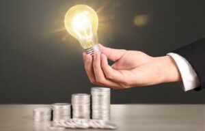 Ręka trzymająca świecącą żarówkę nad stosami monet – symbol oszczędzania energii i niższych rachunków za prąd