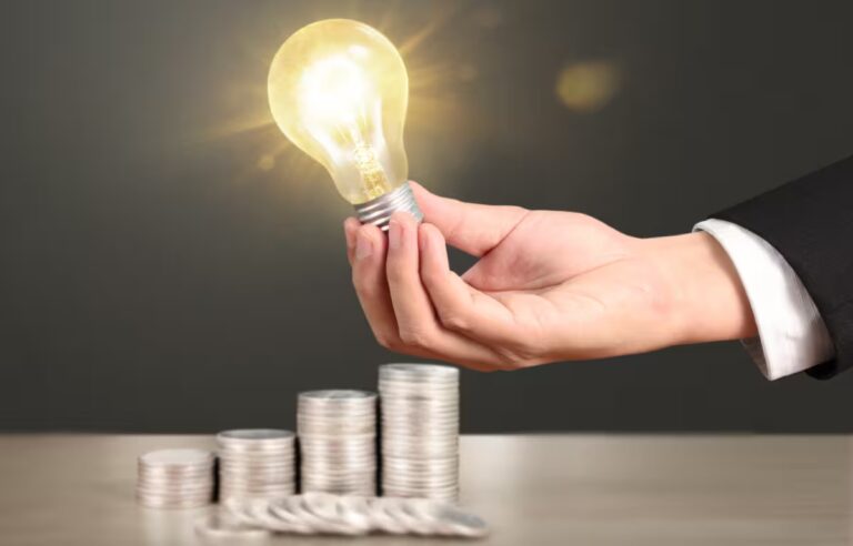 Ręka trzymająca świecącą żarówkę nad stosami monet – symbol oszczędzania energii i niższych rachunków za prąd